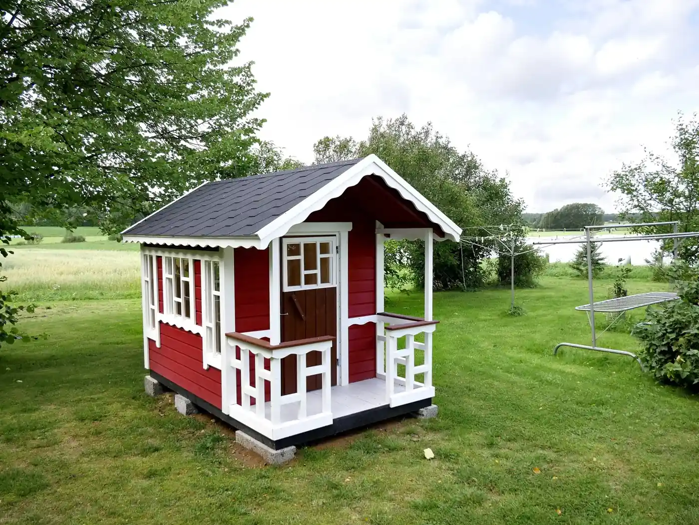 Cabane d'enfant en bois rouge et blanc avec une petite terrasse à balustrade, posée sur des appuis dans un jardin