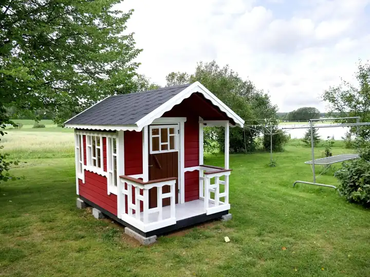 Cabane d'enfant en bois rouge et blanc avec une petite terrasse à balustrade, posée sur des appuis dans un jardin
