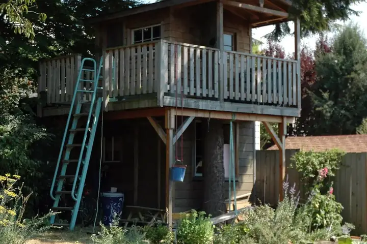Cabane en bois à deux niveaux sur poteaux, avec balcon à garde-corps et échelle